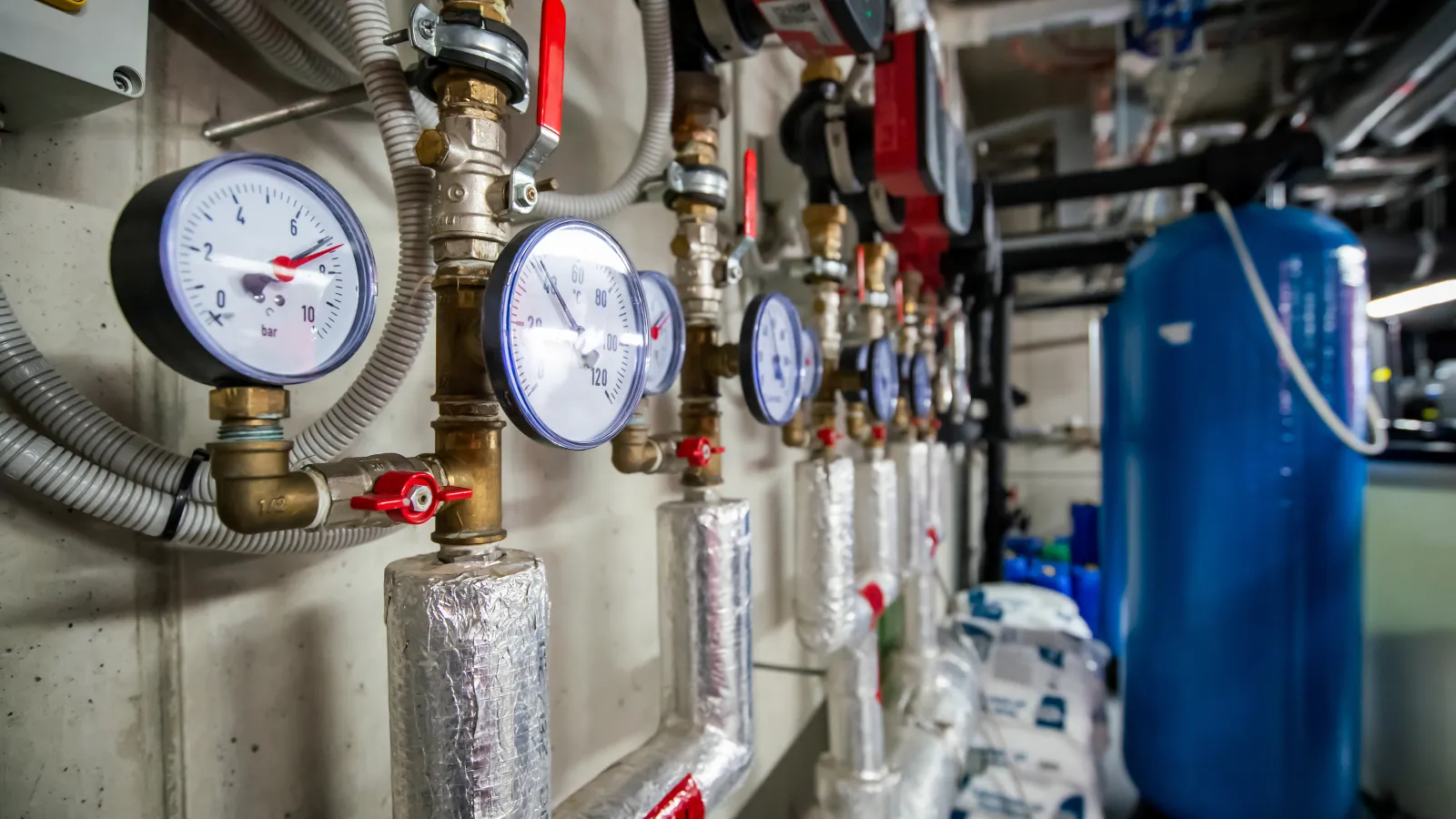 Row of pressure gauges and insulated pipes in a mechanical room with a large blue tank in the background