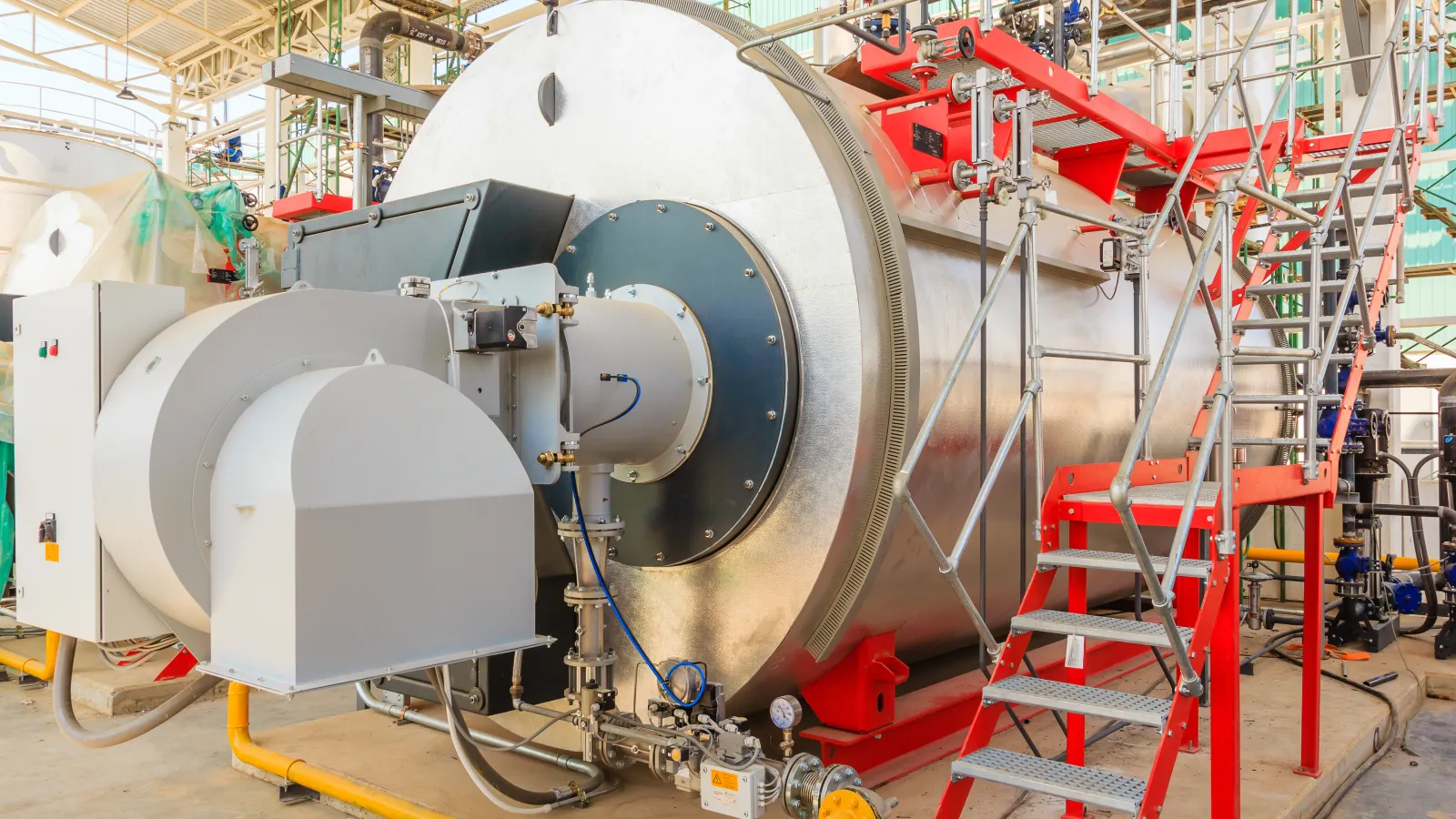 Large industrial boiler system with metal pipes and red safety stairs in a factory setting.