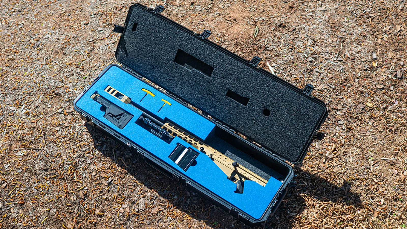 Tactical rifle with accessories neatly placed inside a blue foam-lined black hard case on the ground outdoors.