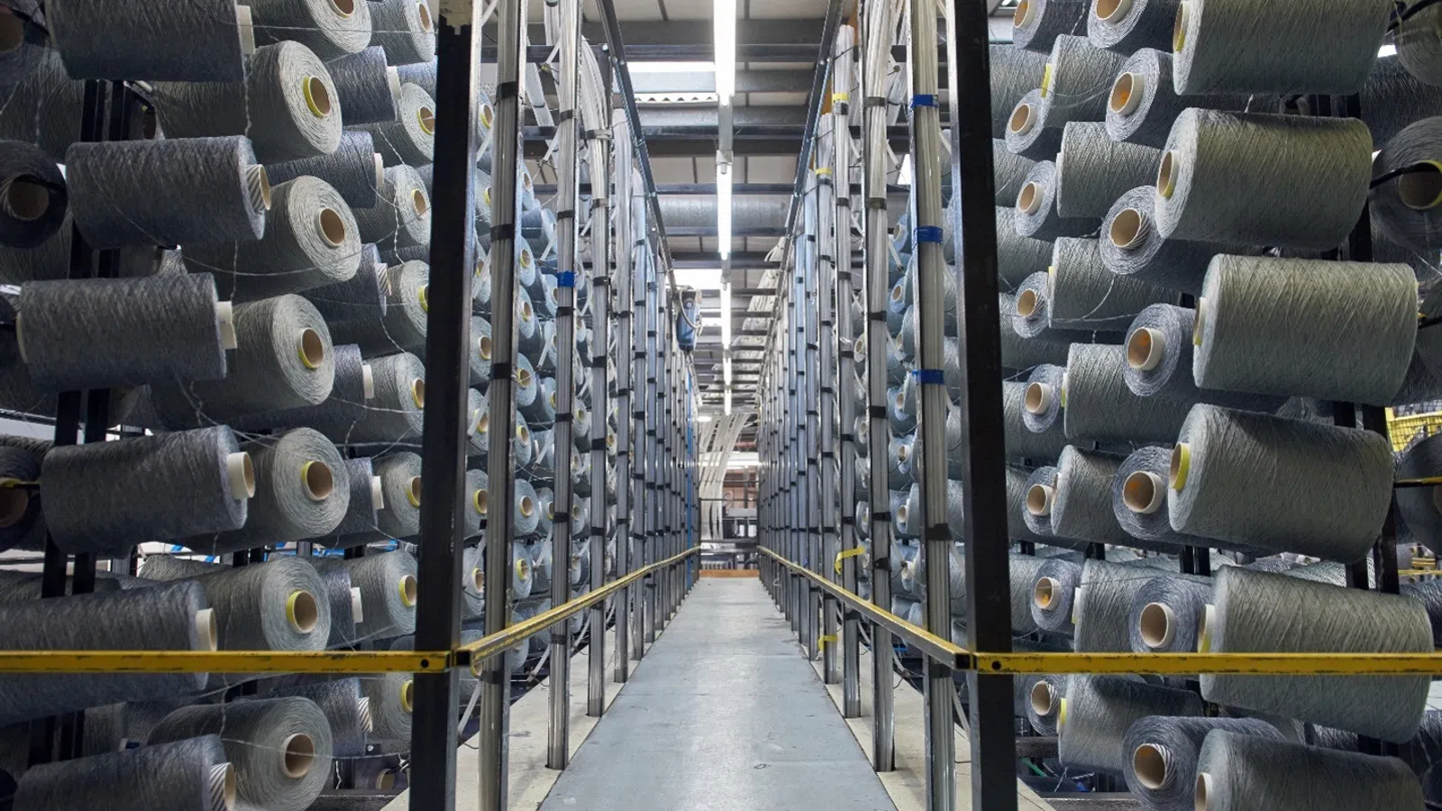 Industrial warehouse aisle lined with large spools of grey textile yarn on both sides under bright overhead lights.