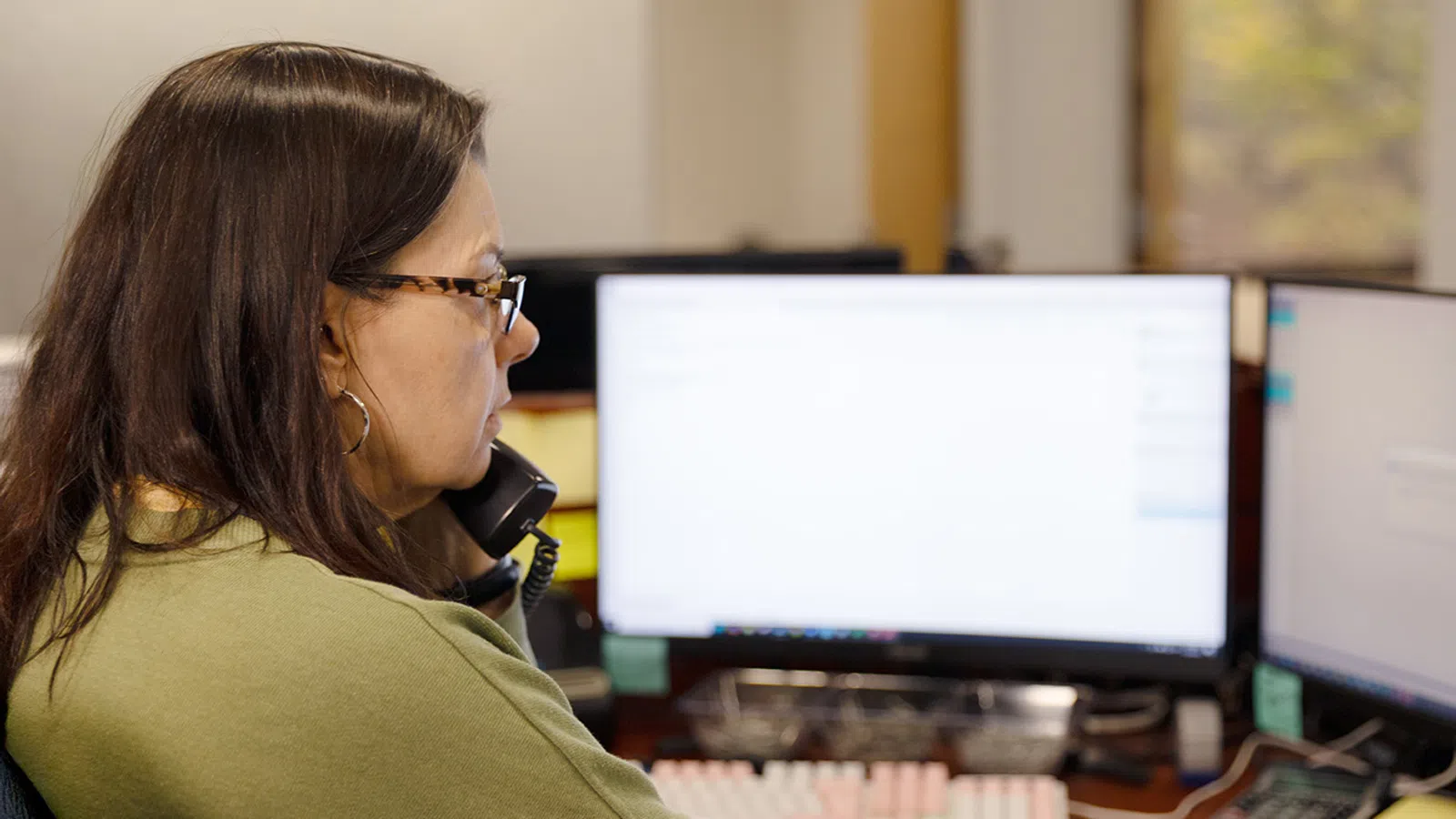Woman in green sweater using computer and phone at office workstation with dual monitors.