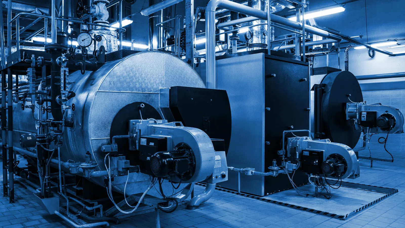 Industrial boilers and complex piping system in a large factory with blue lighting.