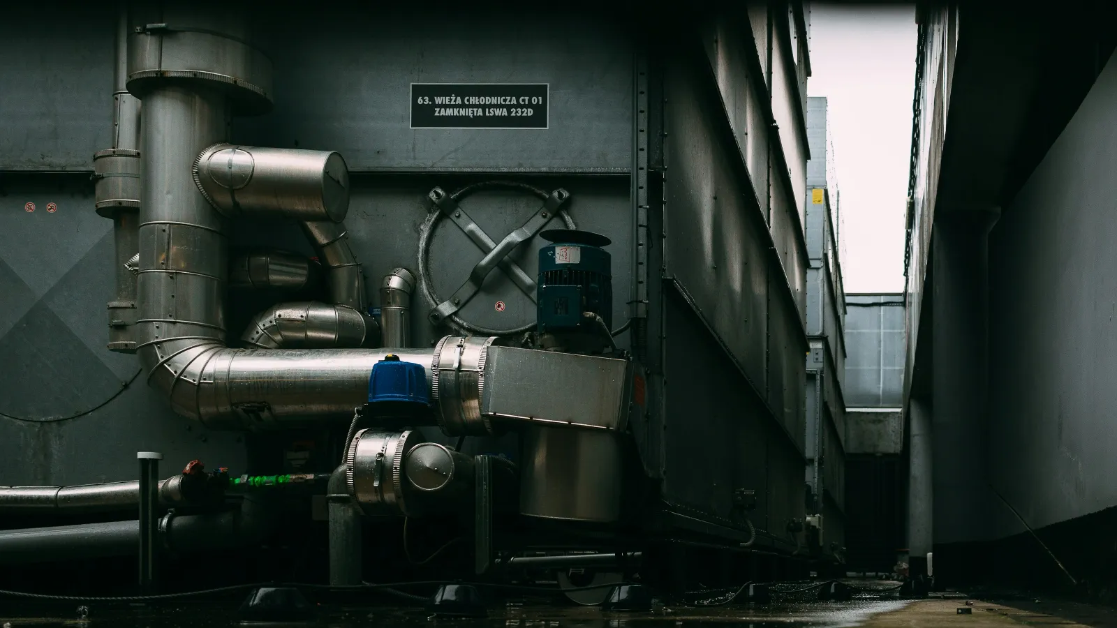 Dark industrial setting with large metallic pipes and machinery against gray walls in a narrow alleyway.