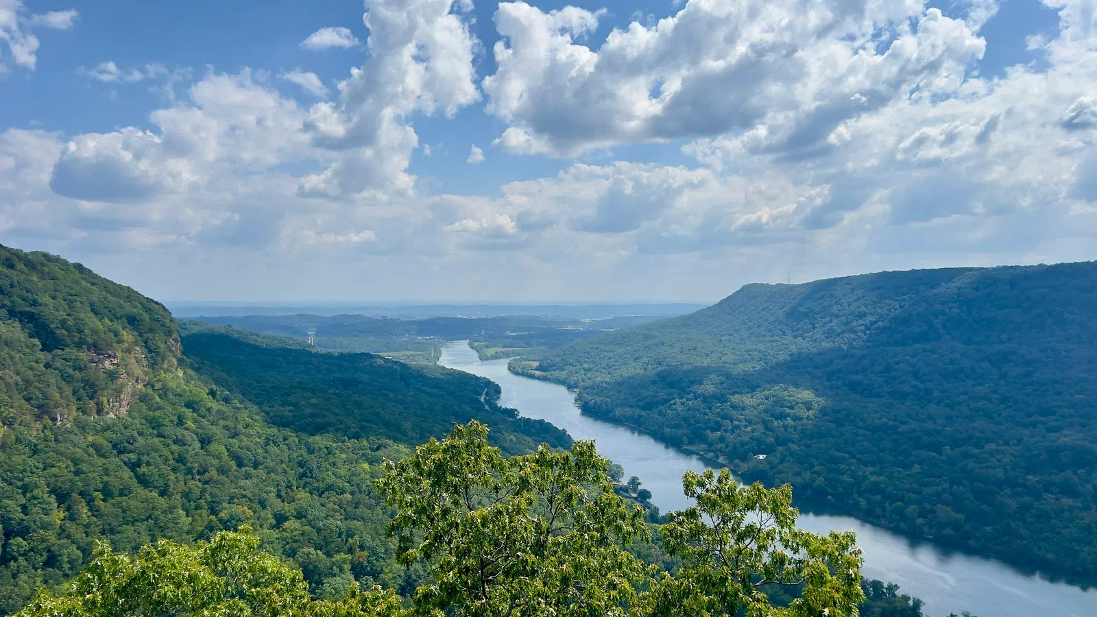 Wide river flowing through lush green forested mountains under a blue sky with scattered clouds