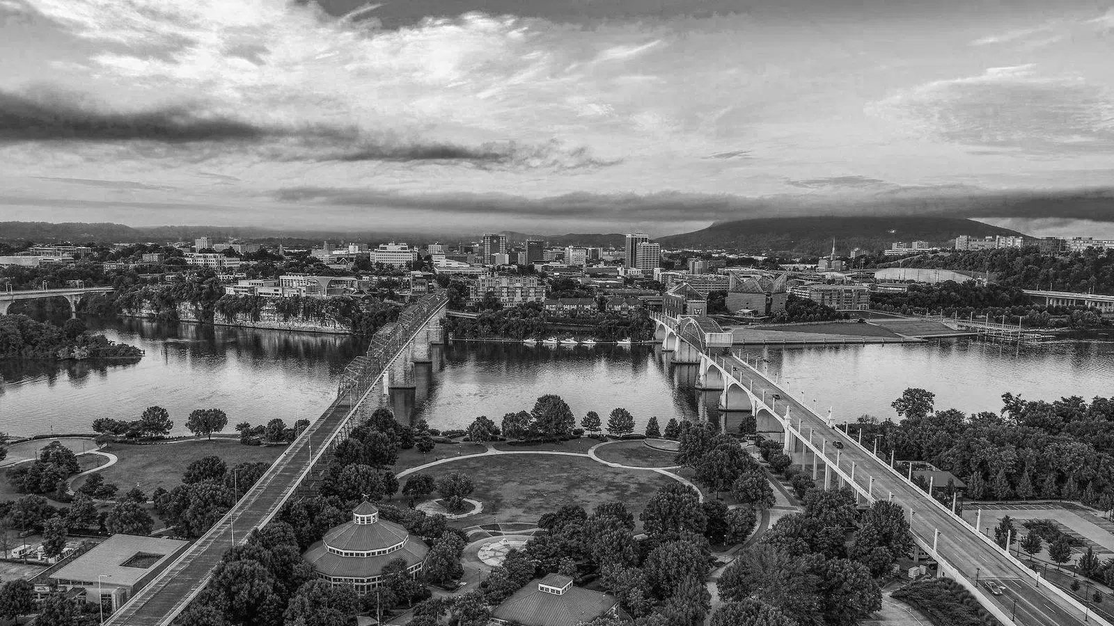 Black and white aerial view of a city riverfront with multiple bridges and urban skyline under cloudy sky.