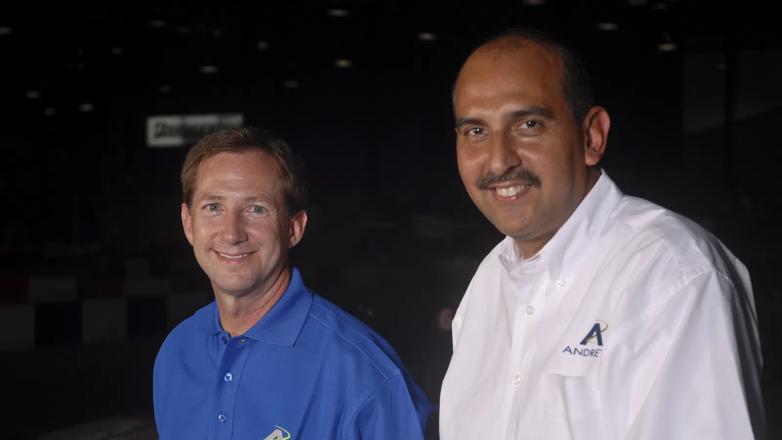 Two men in Andretti branded shirts smiling, one in blue polo and the other in white button-down, dark background.