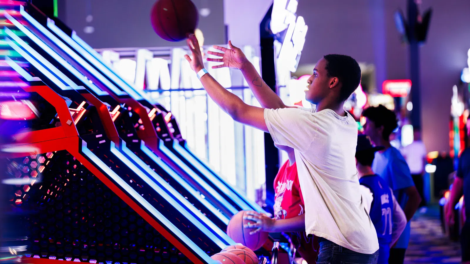 Teen boy playing arcade basketball game aiming for a shot in a brightly lit gaming center.