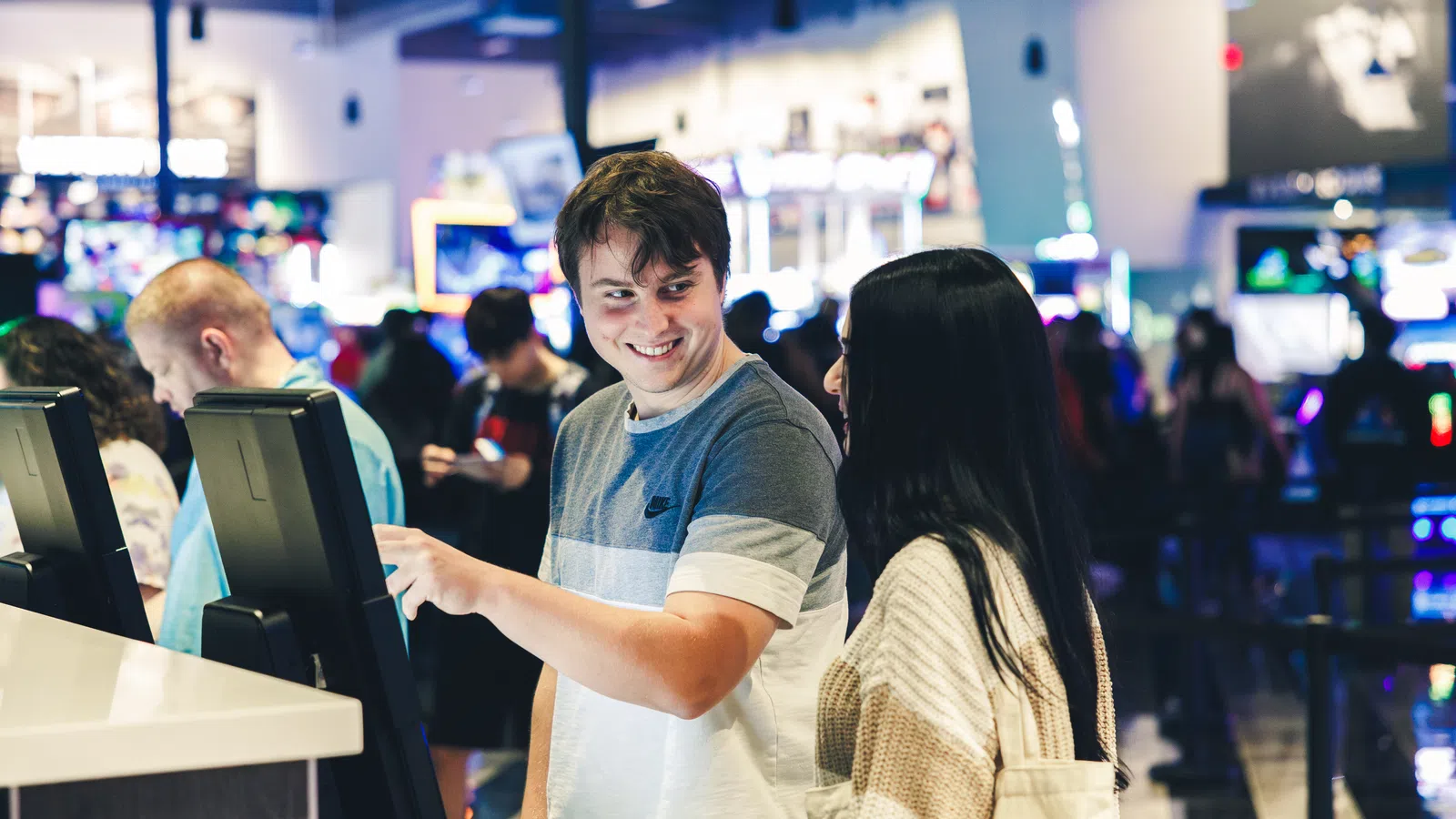 Young man and woman using touchscreen kiosks in a bright, busy arcade or entertainment center.