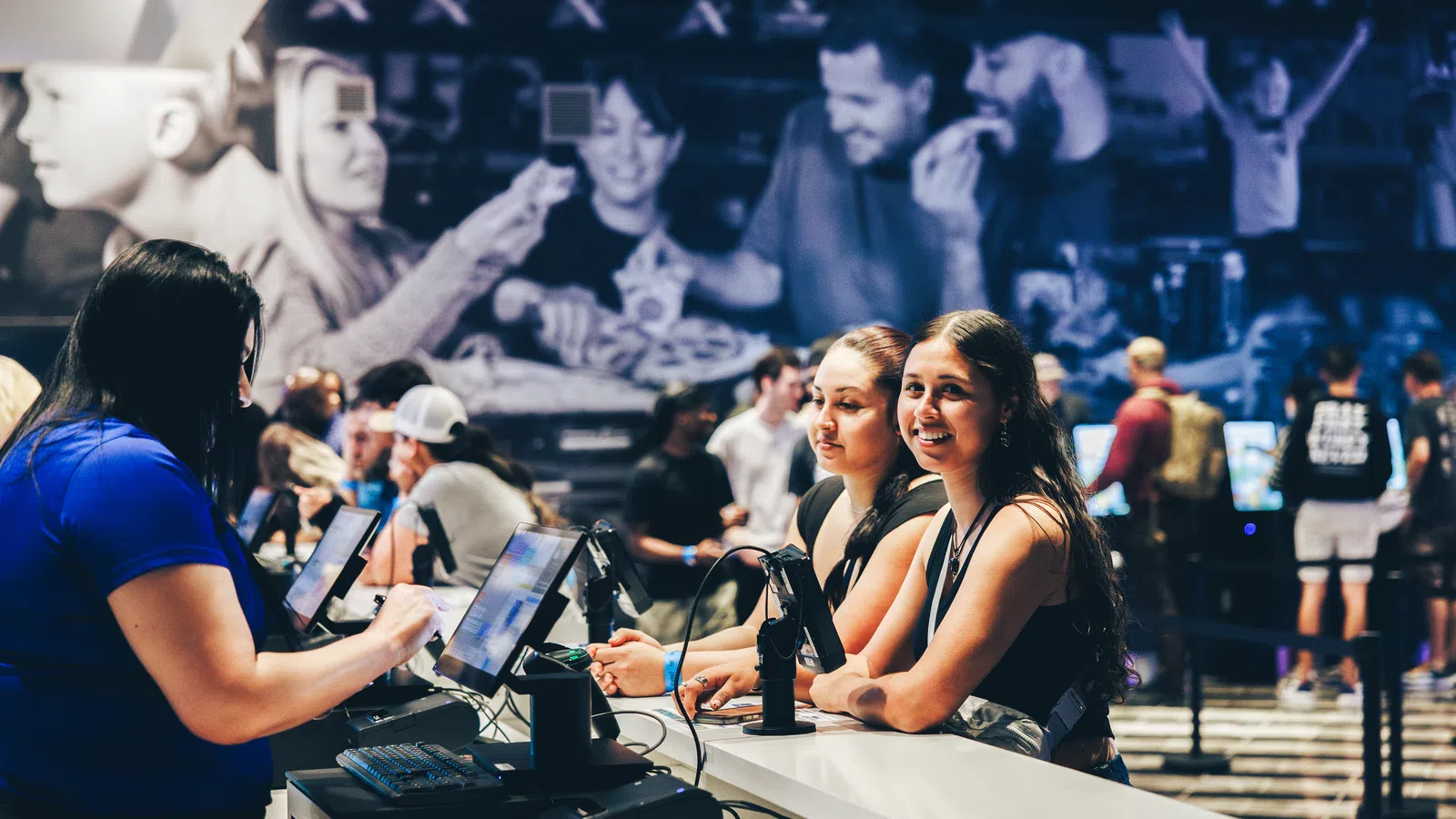 Young women engaging with touchscreen kiosks at a modern, busy indoor entertainment venue with a large mural background