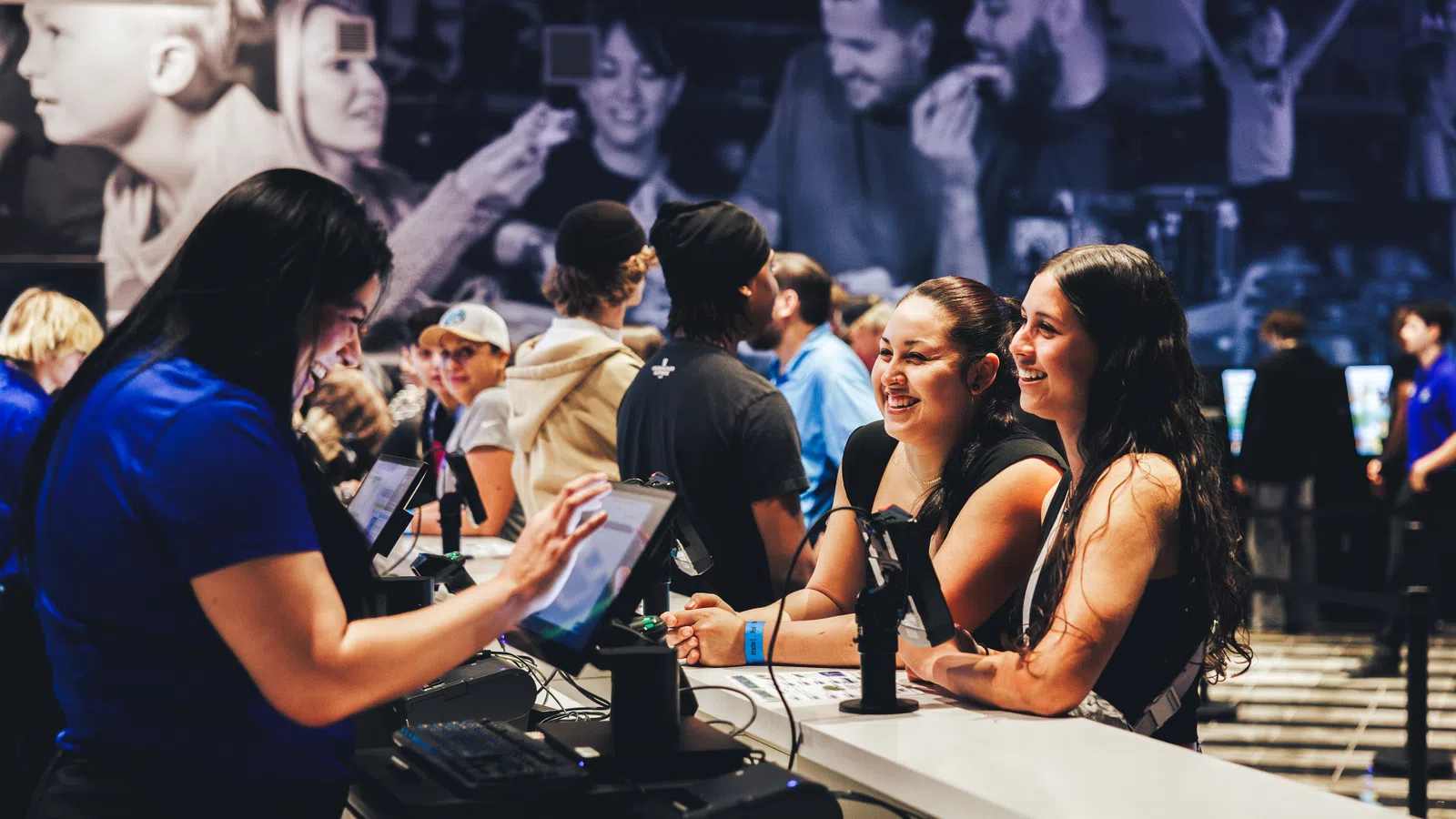 People smiling and interacting at a busy counter with a cashier using touchscreen devices in a vibrant indoor setting