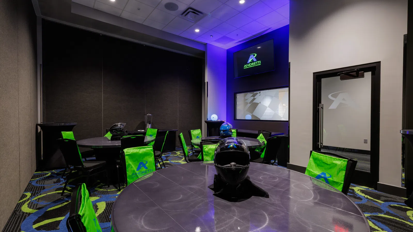 Indoor lounge with black tables, helmets on tables, green Andretti chairs, and screen with overlaid logo text.