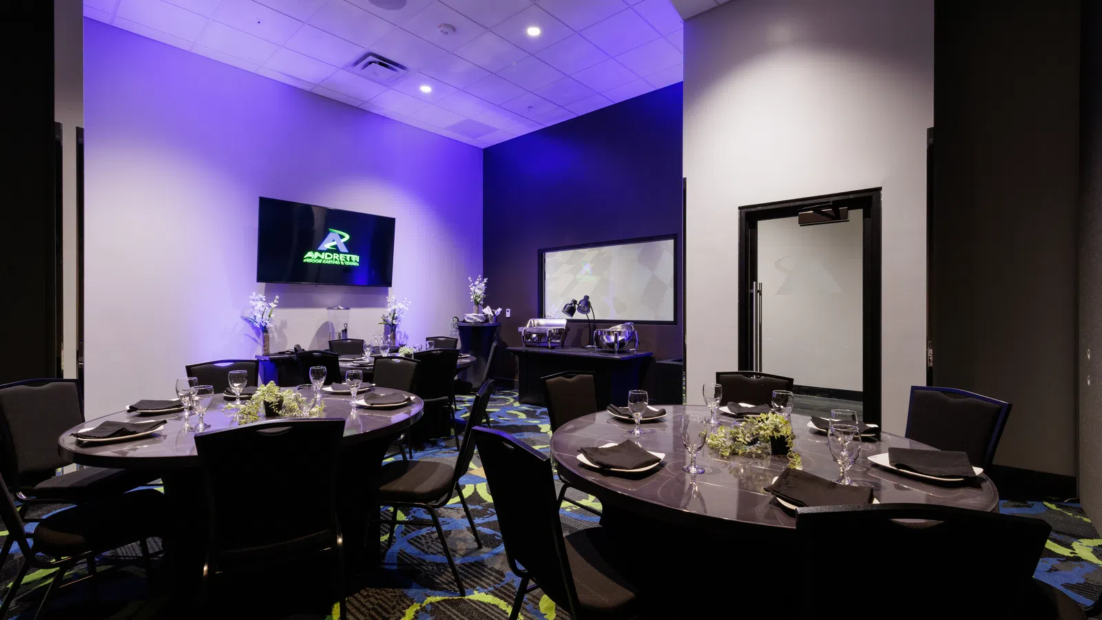 Modern banquet room with round tables, black chairs, elegant table settings, and mood purple lighting.