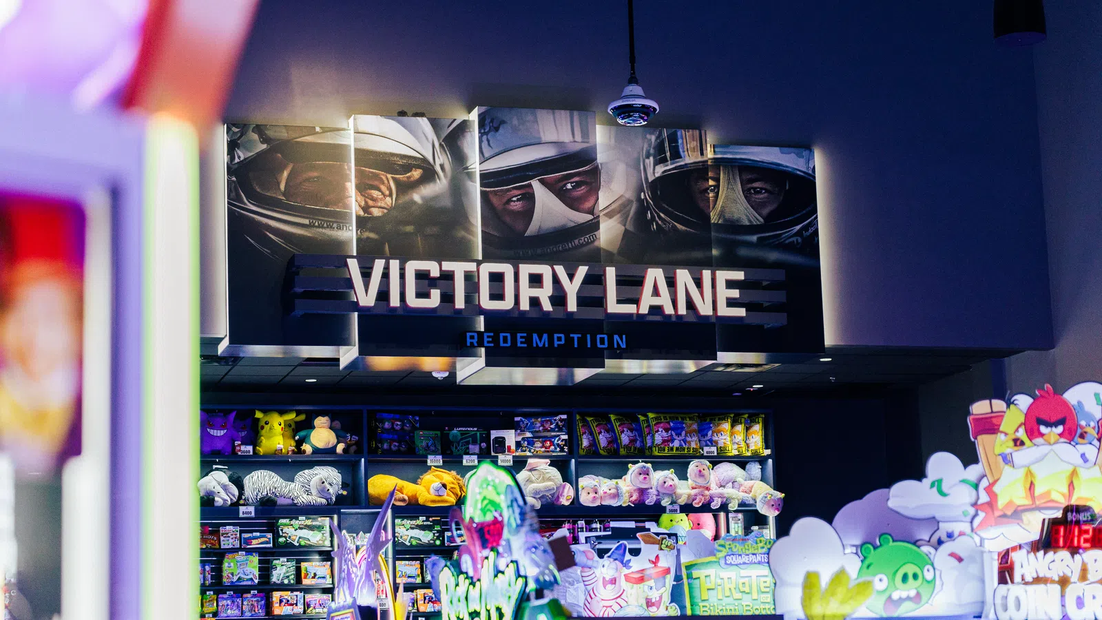 Arcade area with plush toys and game machines under Victory Lane Redemption sign featuring race car driver helmets.