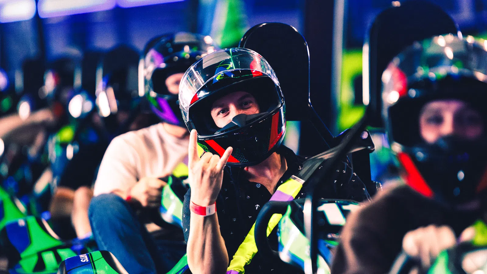 Group of go-kart racers wearing helmets driving indoors, with one racer making a hand gesture.