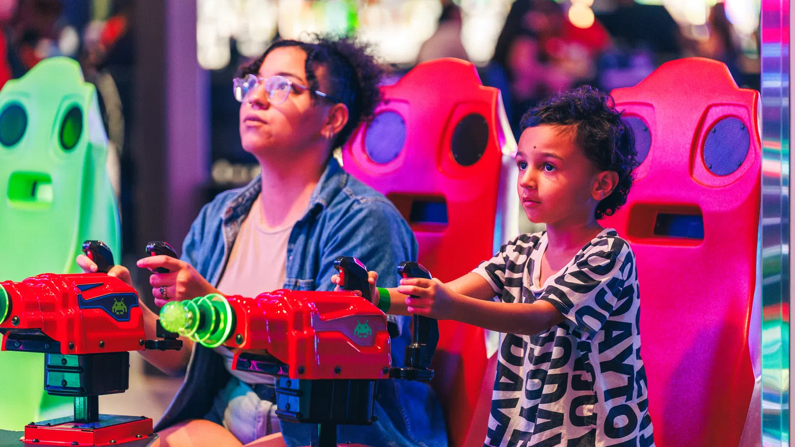 A woman and child playing a vibrant arcade shooting game sitting on colorful red and green seats