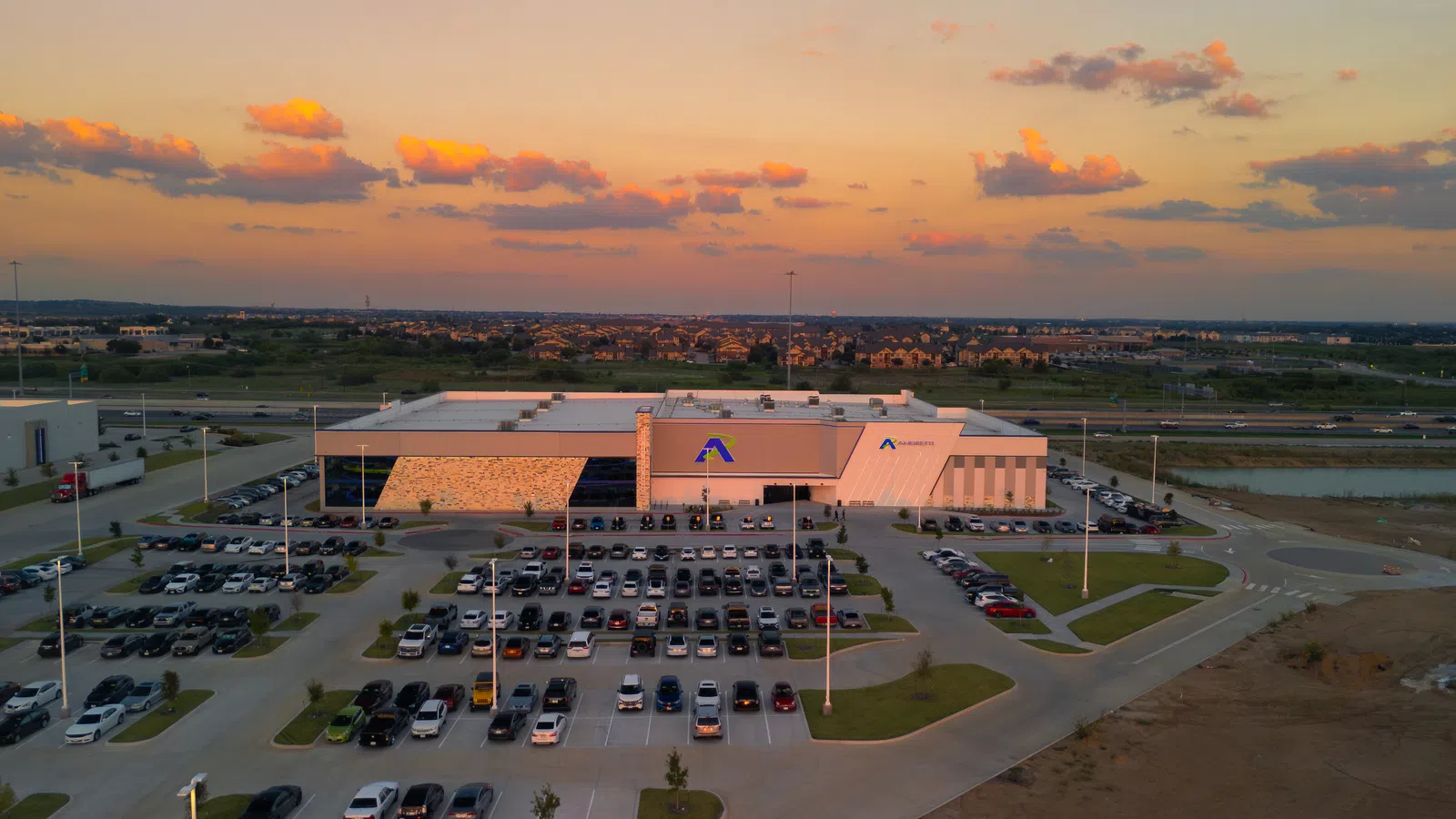 Aerial view of a large industrial building and parking lot at sunset with scattered clouds in the sky.