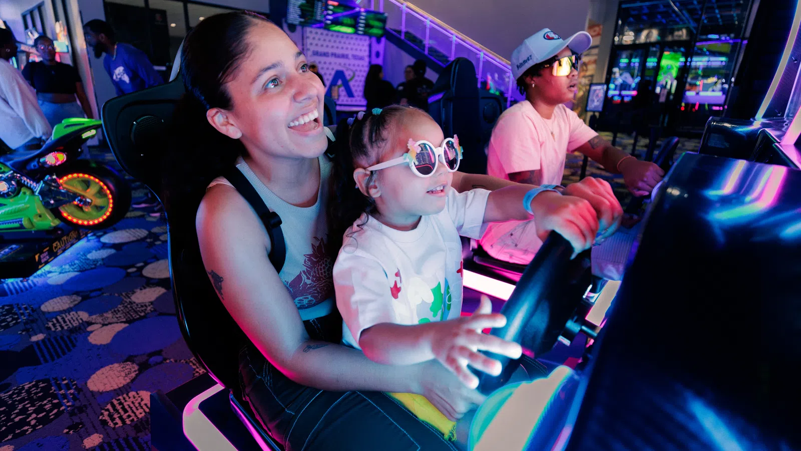 Smiling woman and child wearing sunglasses playing arcade racing game with neon lights in gaming arcade.