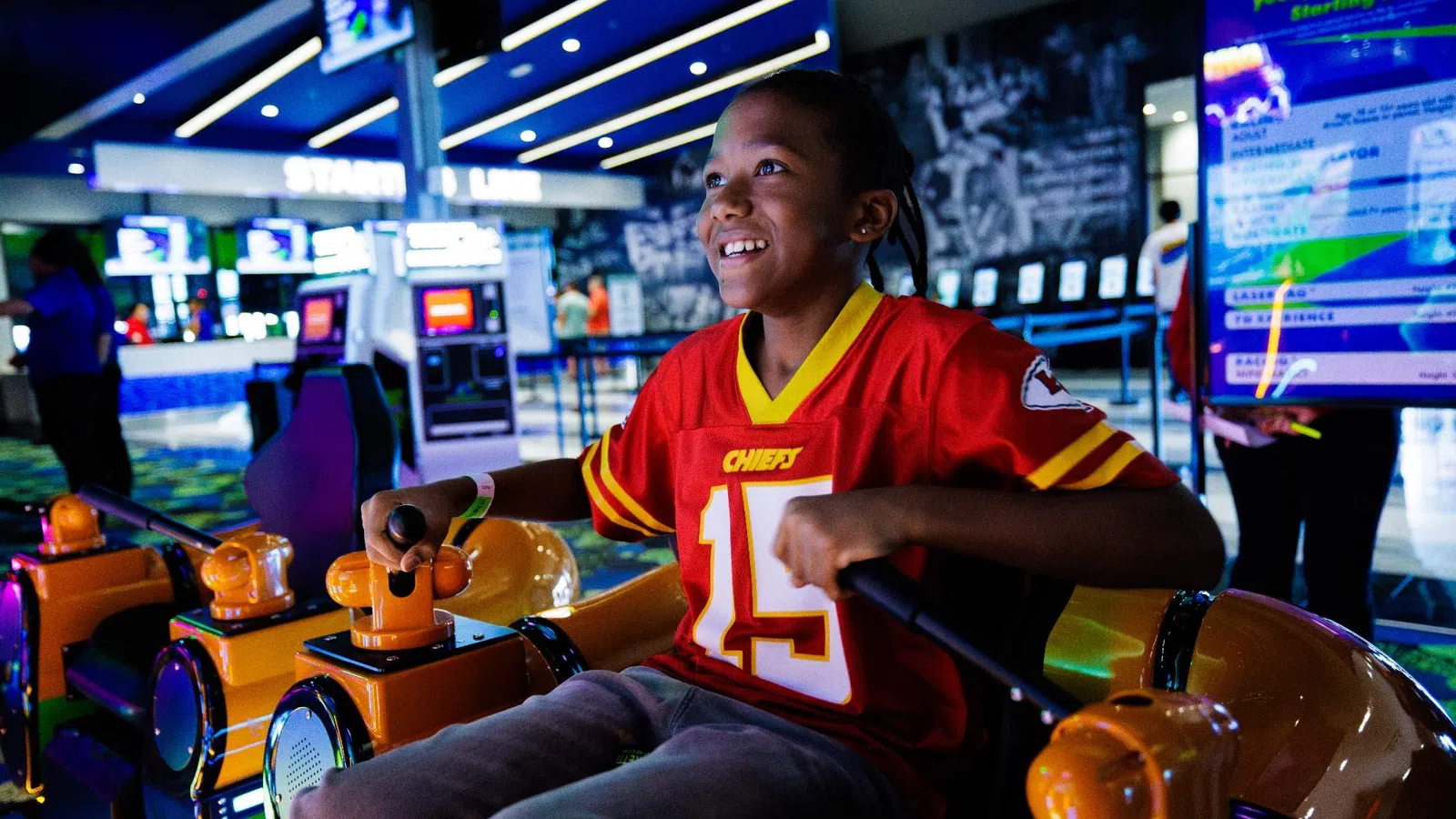 Smiling boy wearing a red Chiefs jersey enjoying a fun arcade game with bright lights and colorful controls.