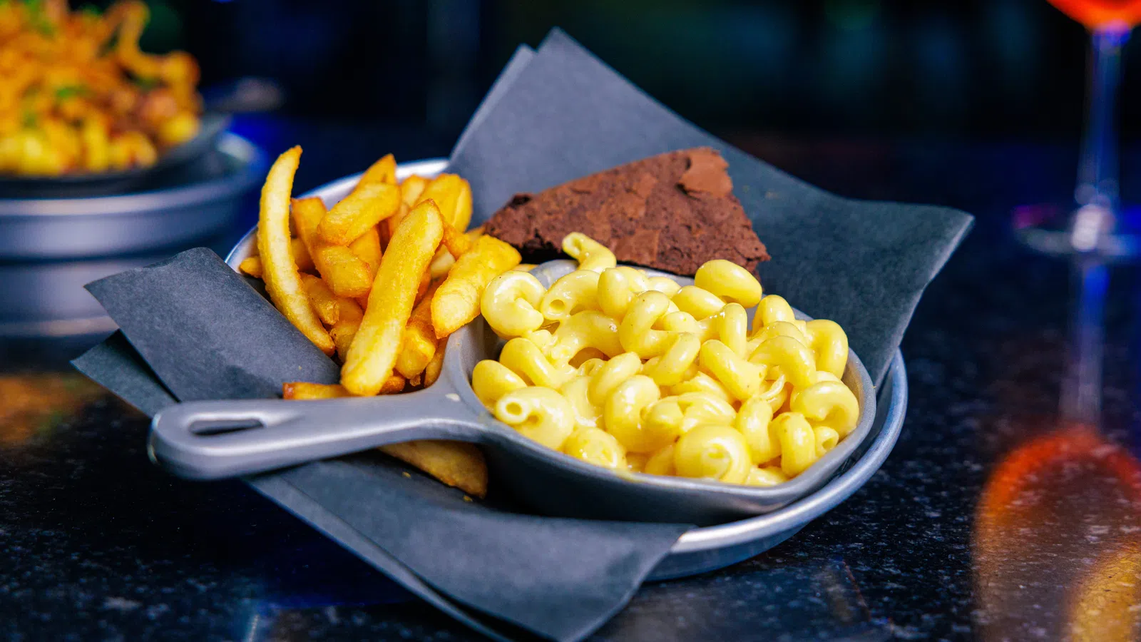 Plate with french fries, creamy macaroni and cheese, and a piece of chocolate brownie on black napkin.