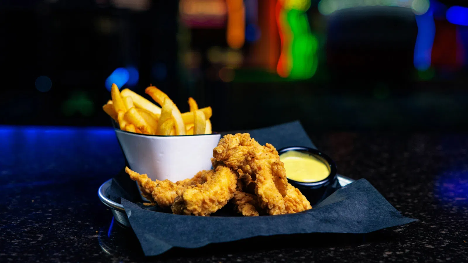Golden fried chicken tenders with crispy fries and dipping sauce on a black tray with colorful blurry background.
