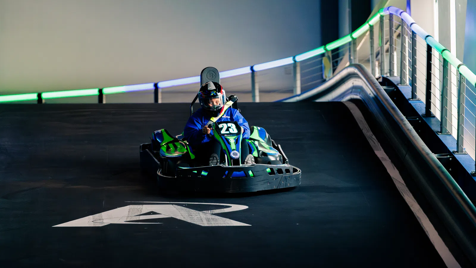 Person wearing helmet driving go-kart number 23 on an illuminated indoor race track.