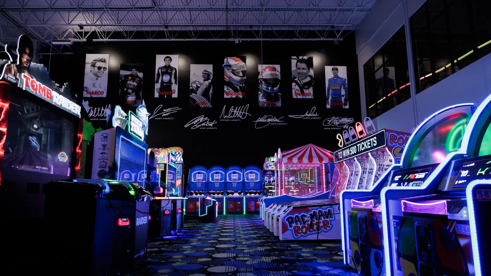 Dimly lit arcade with glowing game machines including Pac-Man, ticket redemption, and racing games under high ceiling.