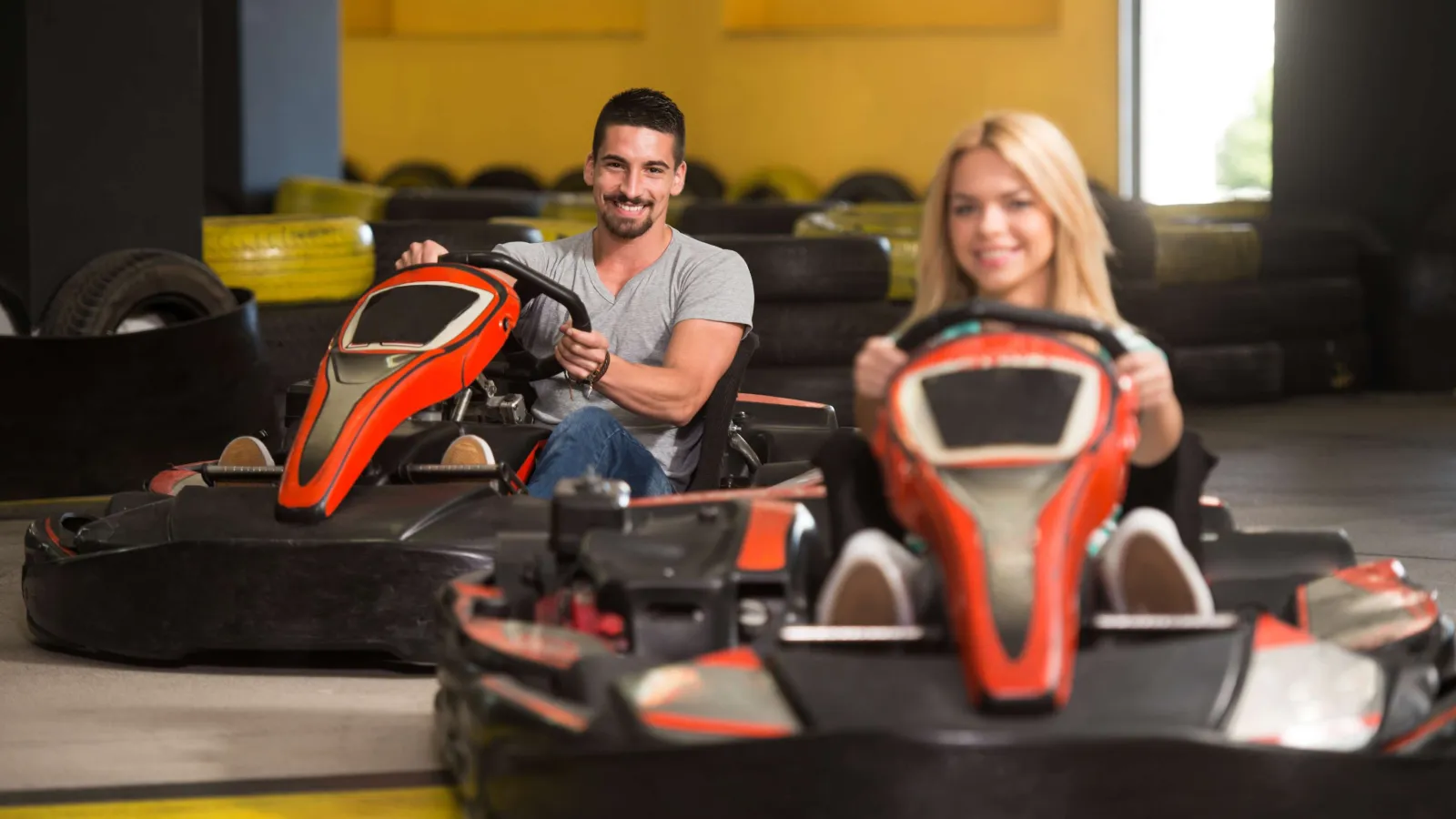 Two smiling adults racing in orange go-karts on an indoor track with tires in the background
