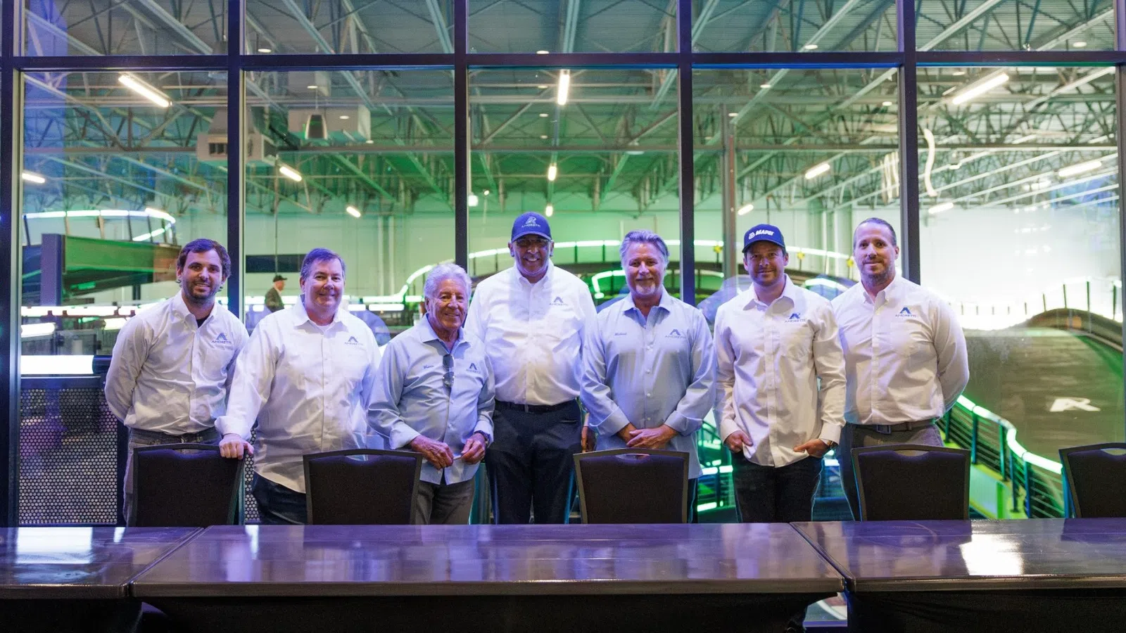 Seven men in light shirts standing behind a large table with a glass wall and industrial setting behind them.