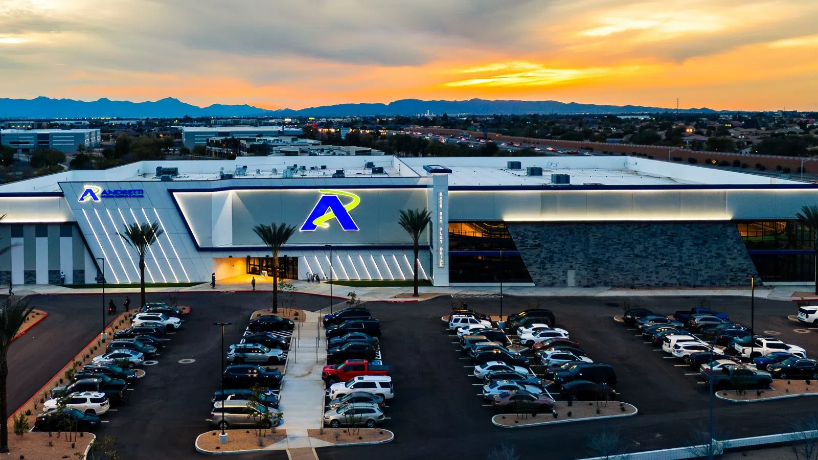 Modern warehouse building with a large parking lot at sunset and mountain range in the background.