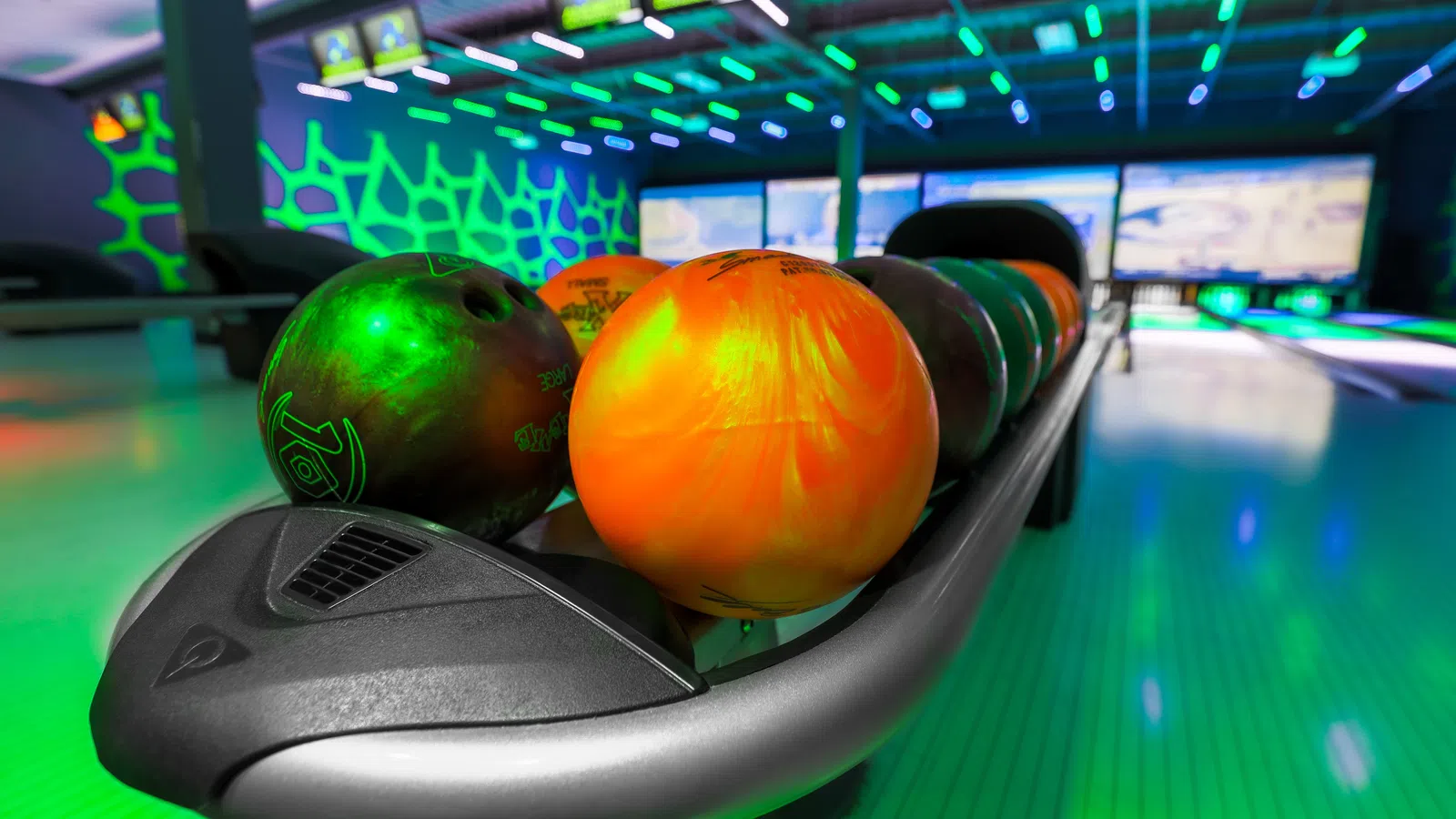 Colorful bowling balls lined up on a sleek rack in a vibrant, illuminated bowling alley.