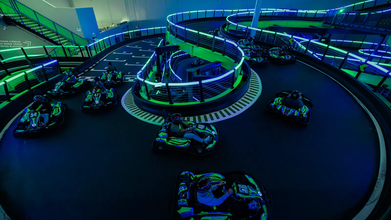 Indoor go-kart track with neon lighting featuring multiple racers navigating a spiral and straight sections of the circuit.