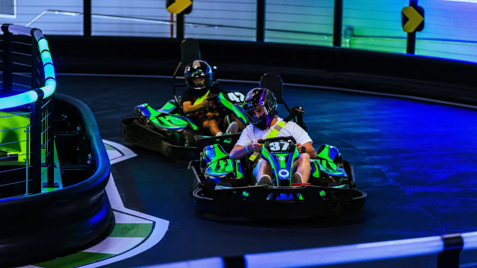 Two people racing green and black go-karts on an indoor track with neon blue and green lights.