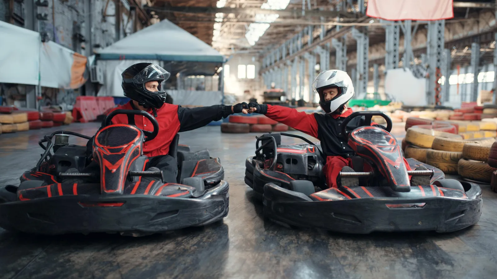 Two people in racing gear fist bump while sitting in go-karts inside an industrial karting arena.