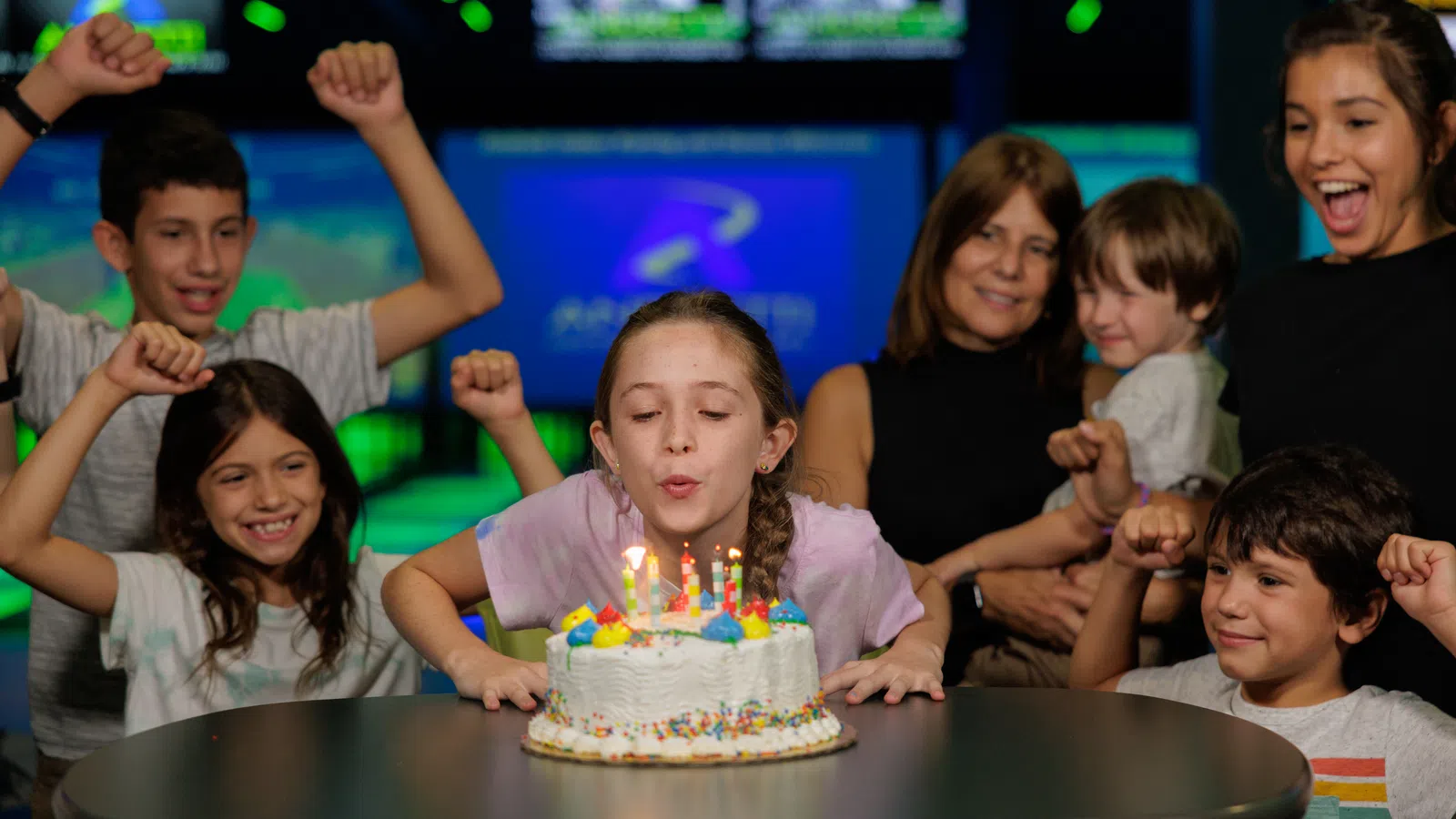 Girl blowing out candles on birthday cake surrounded by cheering family at a bowling alley party.