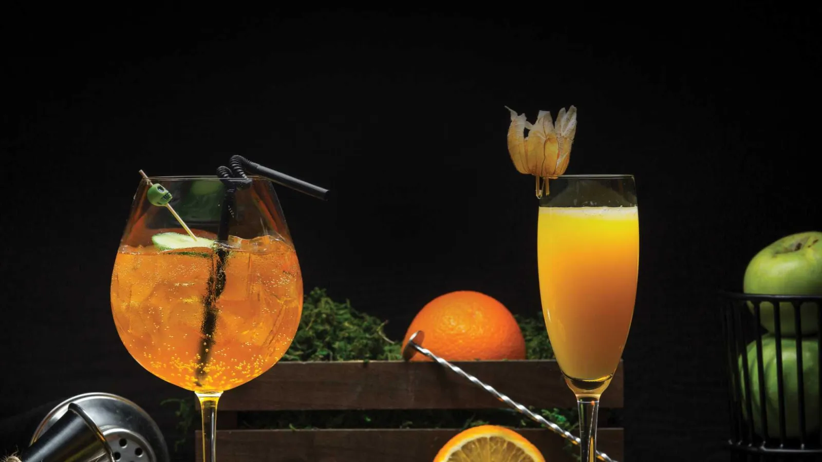 Two vibrant cocktails with citrus garnishes surrounded by fresh oranges, green apples, and herbs on a dark background.