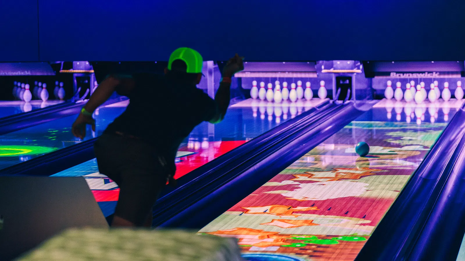 Person wearing neon green hat bowling under blacklight with colorful illuminated lanes and pins ahead