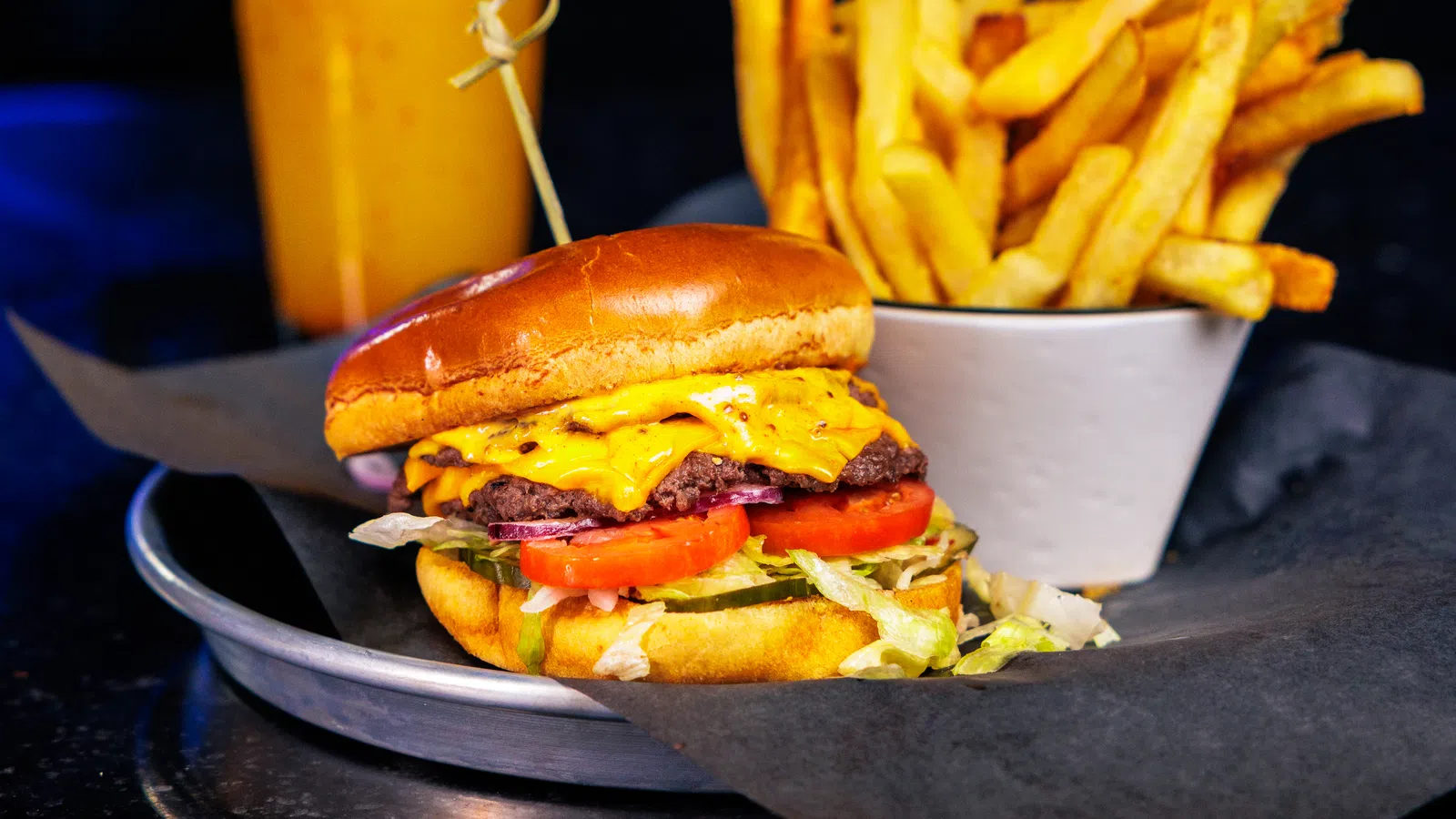 Cheeseburger with lettuce and tomato, cup of French fries, and a drink with colorful lights in background