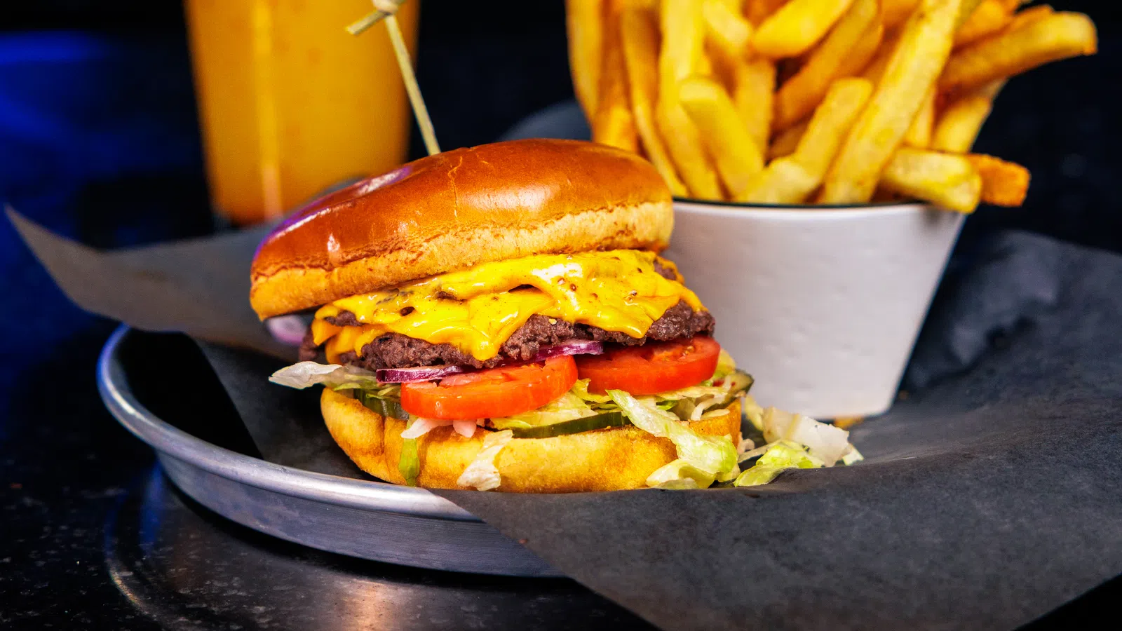 Cheeseburger with lettuce and tomato, cup of French fries, and a drink with colorful lights in background