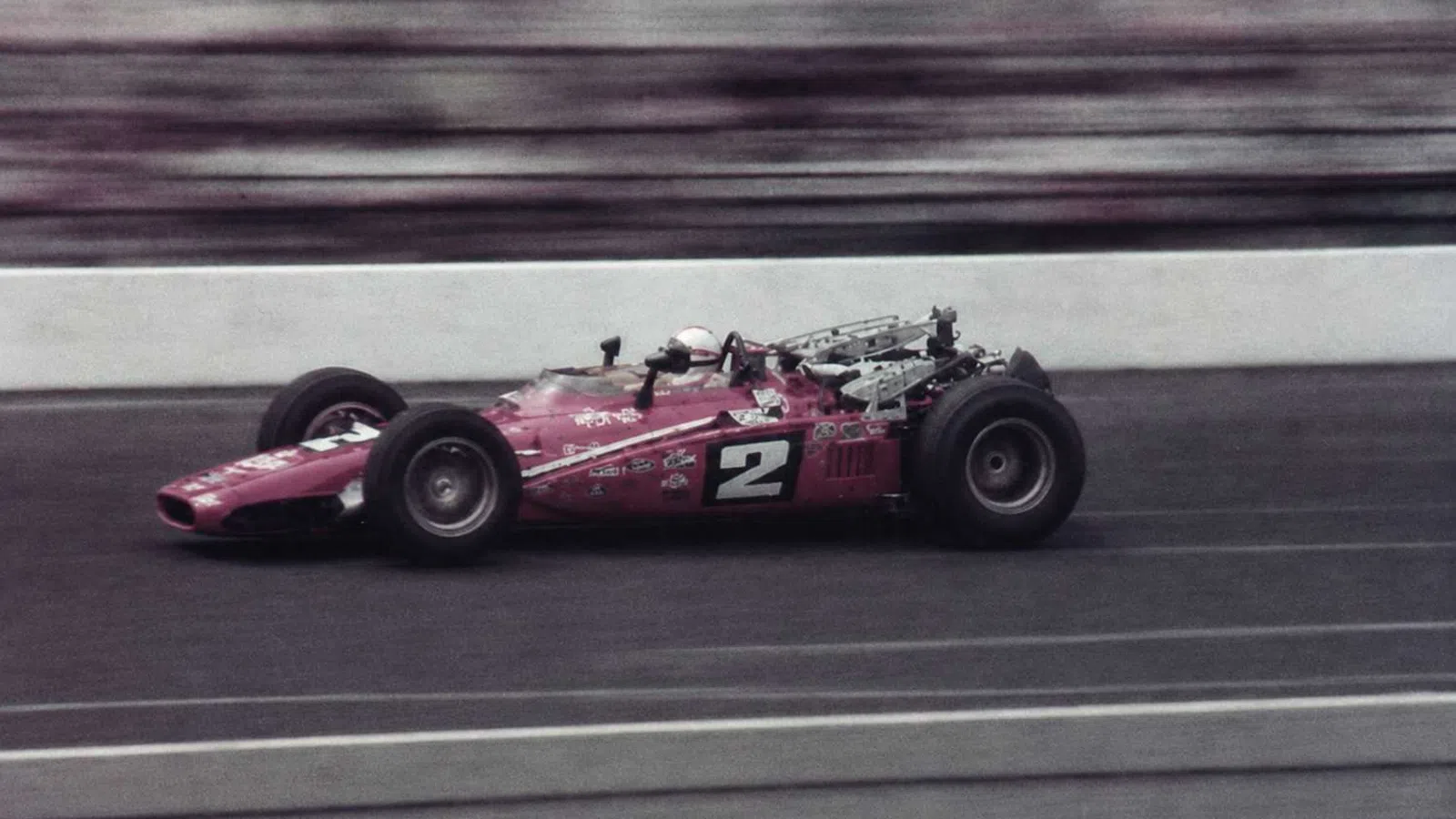 Vintage open-wheel race car number 2 speeding on a racetrack with blurred background and driver in helmet.