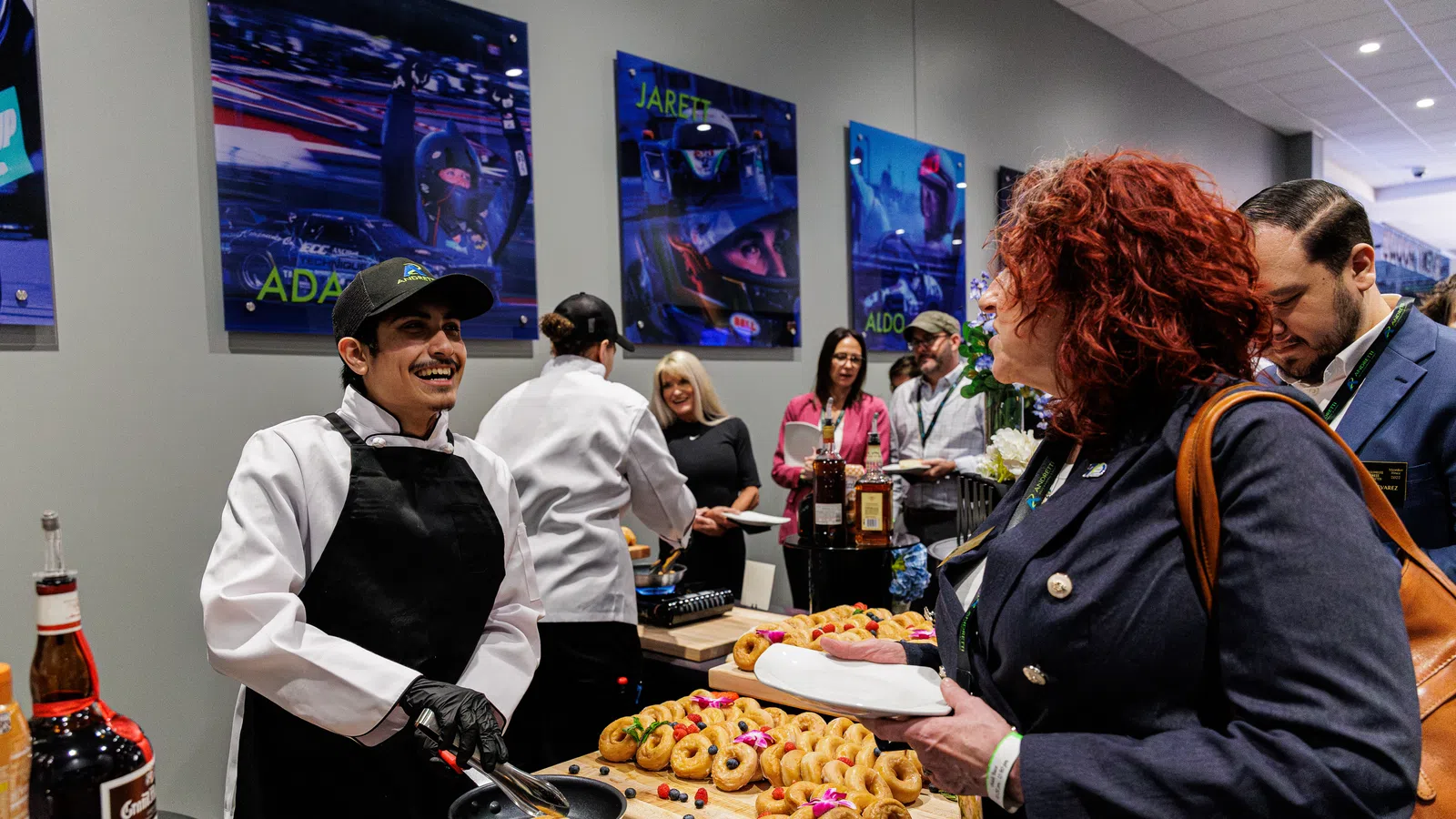 Chef wearing black apron serving pastries to guests at an indoor event with vibrant wall art and refreshments.
