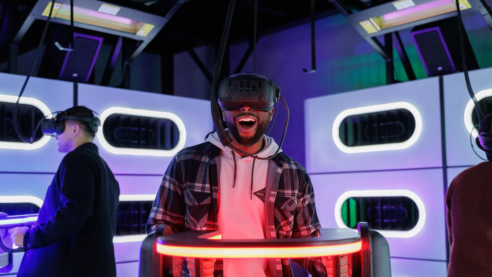 Man wearing virtual reality headset enjoying immersive VR game in neon-lit gaming room with other players.