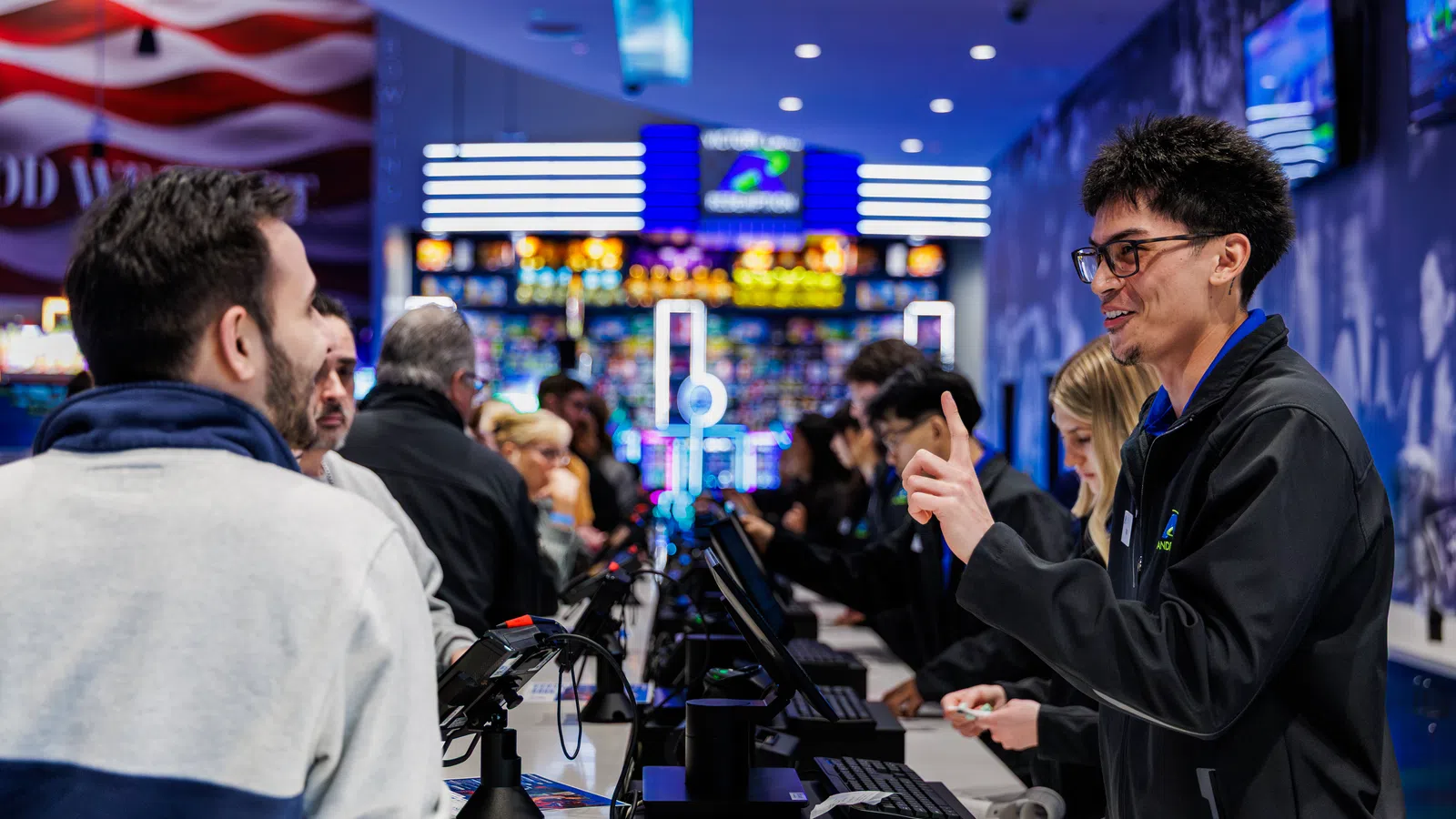 Customers interacting with friendly staff across a bright, busy casino or entertainment venue counter with digital screens.