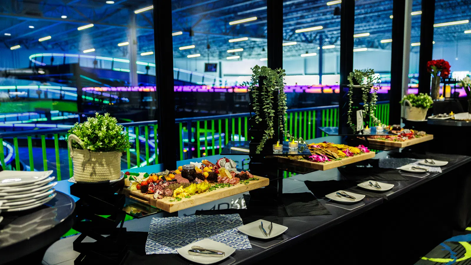 Buffet table with charcuterie boards, plates, and decorative plants inside a modern venue with large windows and vibrant lighting.