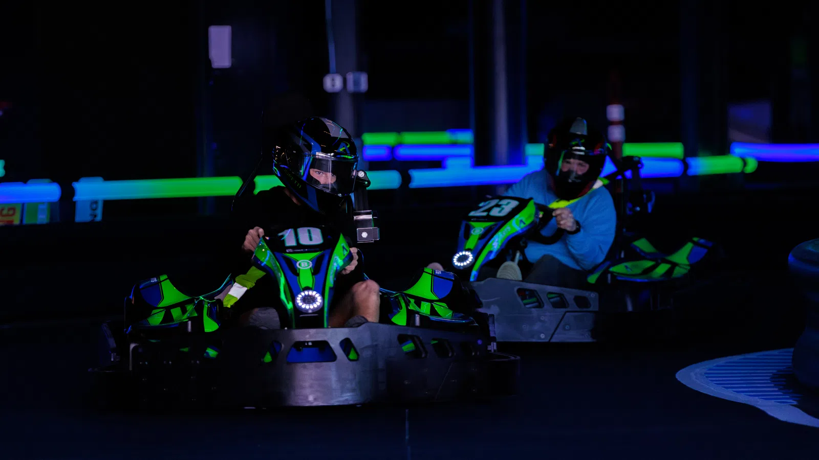 Two adults racing in green and blue glow-in-the-dark go-karts at an indoor track with neon lighting.