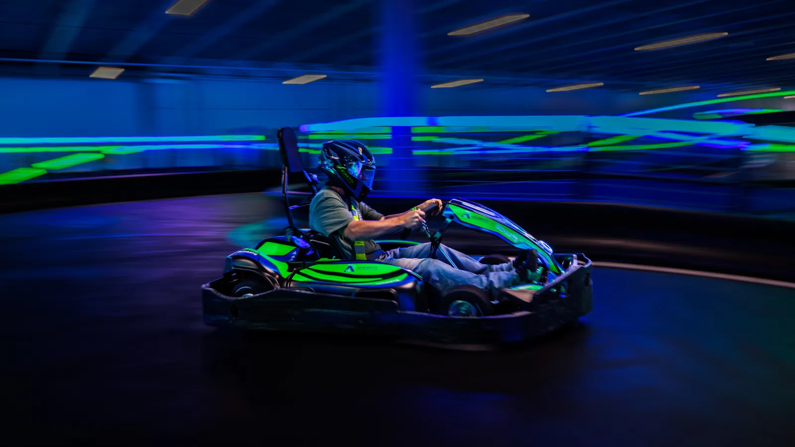 Person driving a green go-kart at high speed on an indoor track with blue neon lights