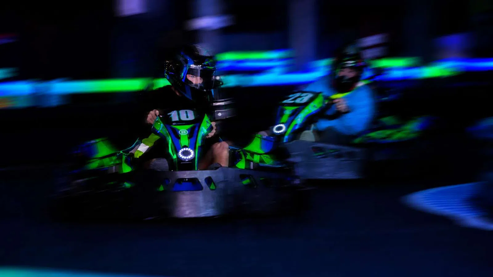 Two racers in green and black go-karts speeding on an indoor track with neon blue and green lighting.