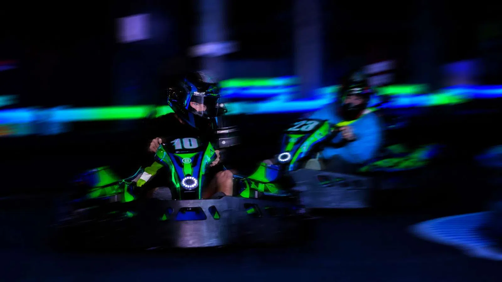 Two racers in green and black go-karts speeding on an indoor track with neon blue and green lighting.