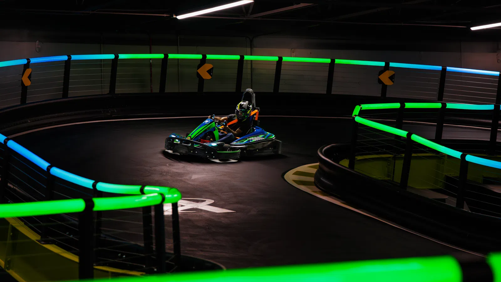Person driving a go-kart on an indoor neon-lit track with green and blue lights and safety barriers.