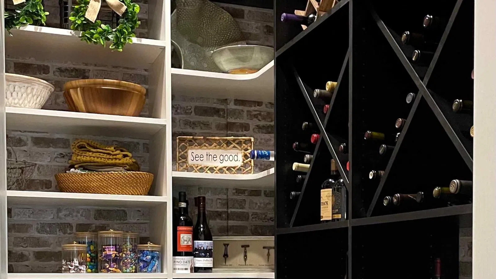 Organized pantry with shelves, baskets, wine racks, drawers, and decorative signs on a brick wall background