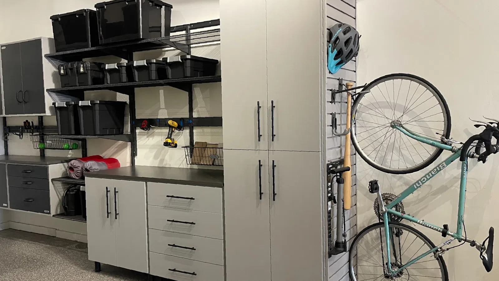 Organized garage with gray storage cabinets, black bins on shelves, and a vertical bike rack holding a teal bicycle and helmet.